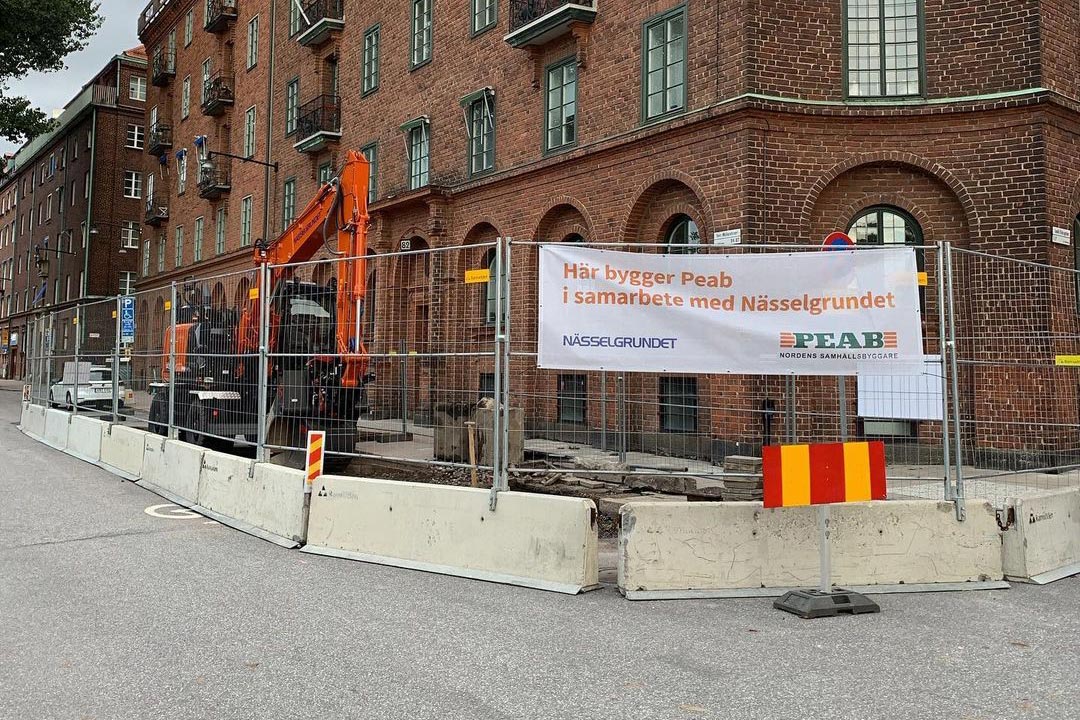Byggarbetsplats med skylt med PEAB och Nässelgrundet logotyper. Tegelbyggnad i bakgrunden.